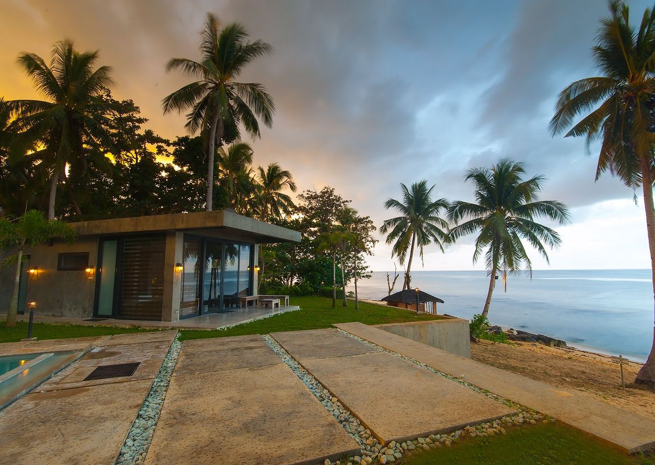 A Shoreline cabin at sunset, concrete and glass with palms and a path to the sea