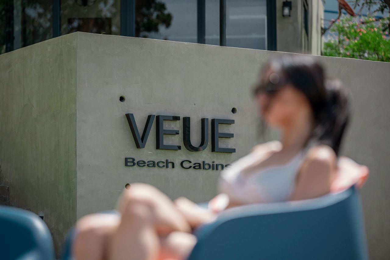 The VEUE Beach Cabins brand mark on a poured-concrete entry wall, with a guest reclining in a teal lounger
