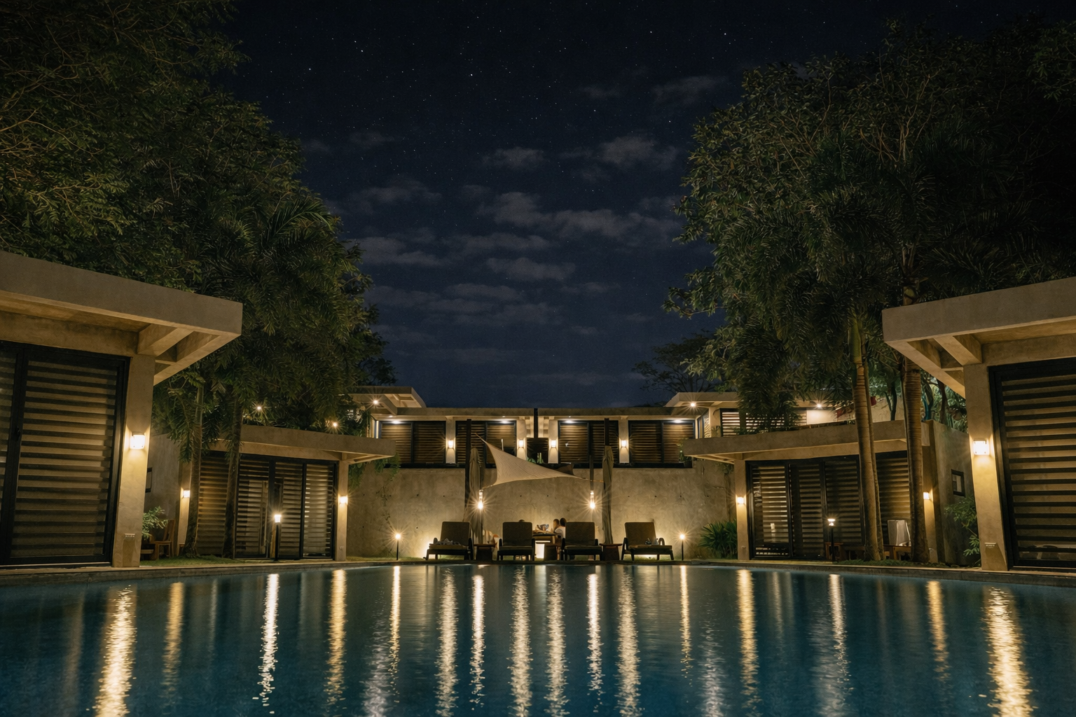 Veue Beach Cabins at night — private cabins on Patar Beach flanking a long courtyard pool, warm pendant lights reflected in the water under a starry sky.