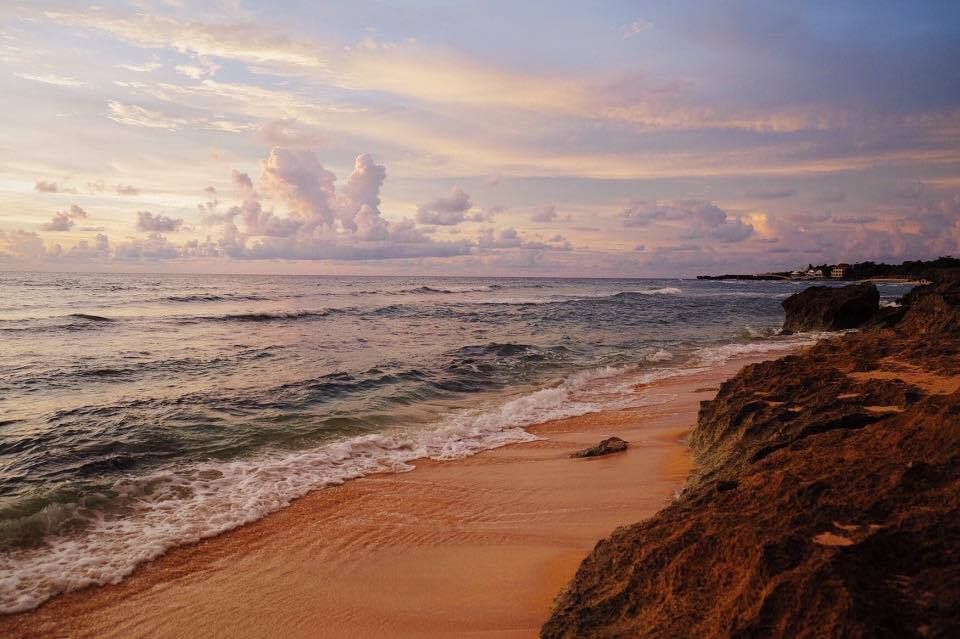 Patar Beach at dusk, calm turquoise water along a long curve of golden sand, with the sky over the West Philippine Sea turning rose and amber.
